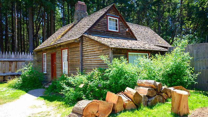 A log cabin with a wooden fence at the back and a pile of large logs in front of it