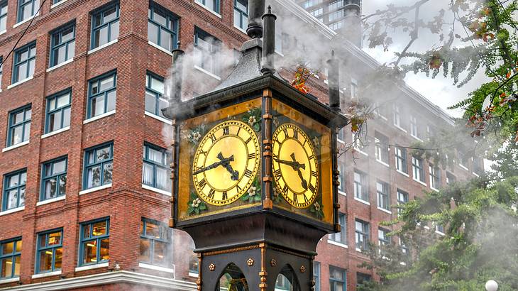 A close up of an old steam clock surrounded by lights, a building and trees