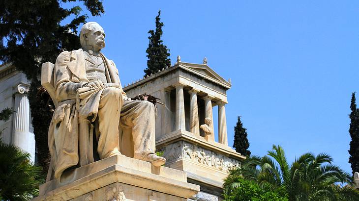 A statue of a seated person next to a building with columns with trees at the back