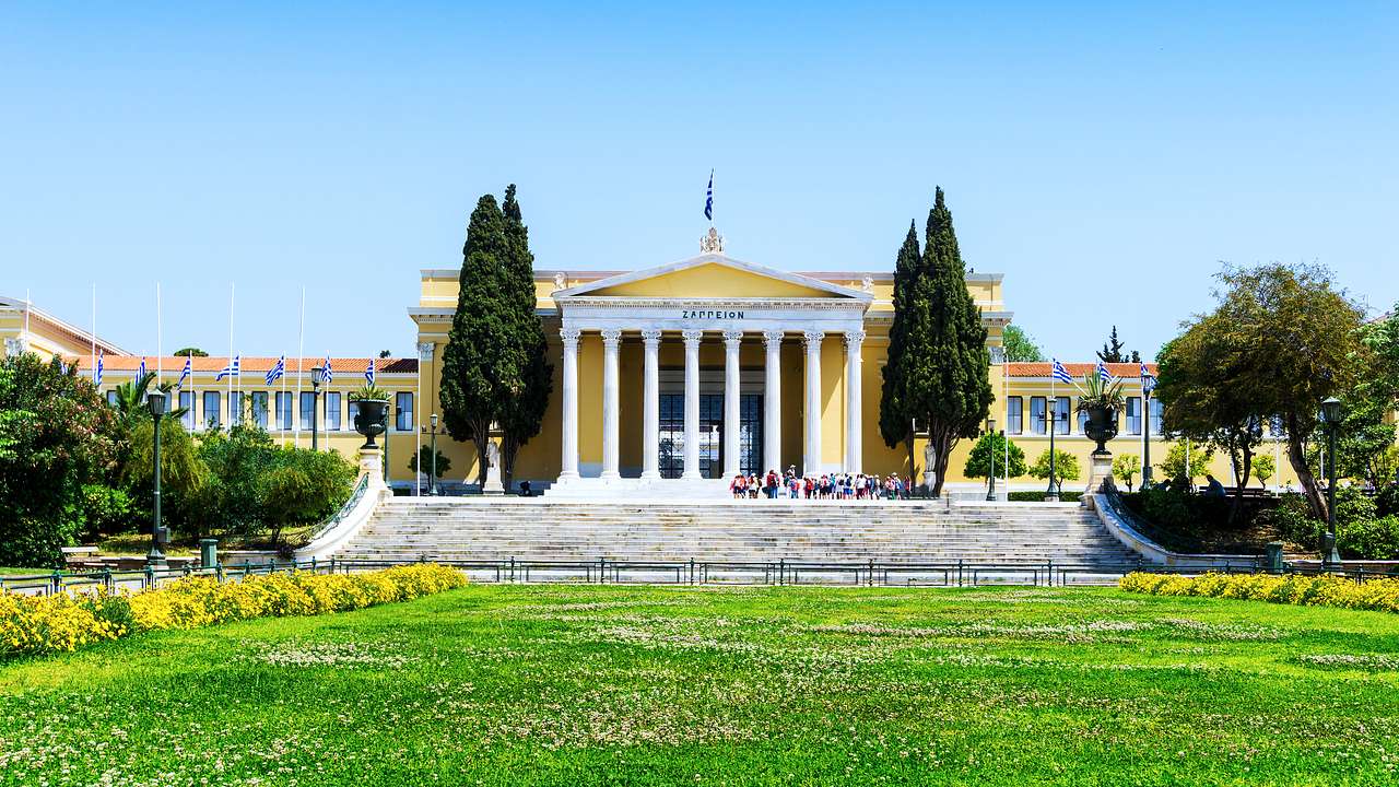 A grand museum with columns and four statues on top and trees and greenery in front