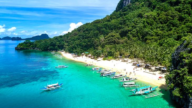 Aerial view of turquoise water, people on sand, trees, and boats moored in water