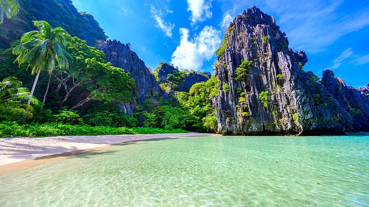 A sandy beach with greenery and mountains in the back and a steep limestone cliff