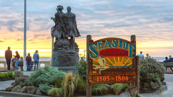 A sign that says Seaside, Oregon, next to a garden and statue by the coast at sunset