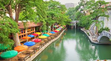 A river with a bridge on one side and trees and colorful patio umbrellas on the other
