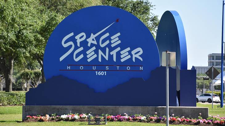 A blue sign that says "Space Center Houston" with grass and greenery around it