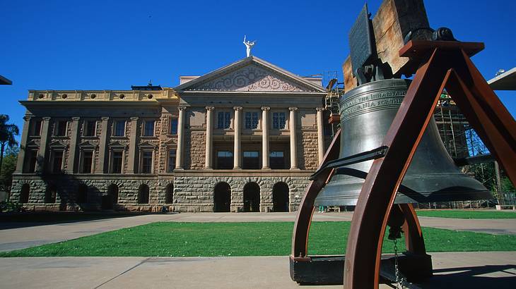 A bell next to a grassy patch and a large building and a walkway