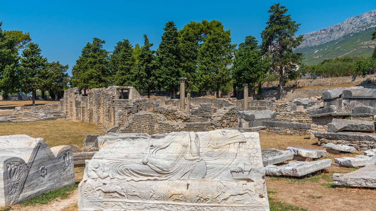 Stone ruins on the grass next to trees, a mountain, and a clear blue sky