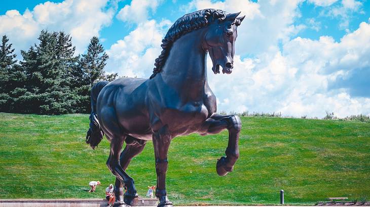 A large statue of a horse next to grass and trees under a blue sky with clouds