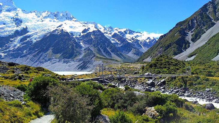 A rocky, grassy hiking trail with a bridge going to snow-capped mountains at the back