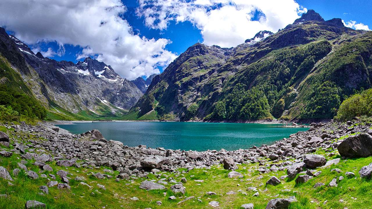 A beautiful blue lake with mountains at the back and grass and rocks in front