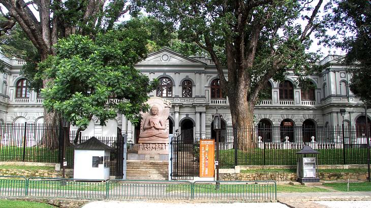 A Buddha statue facing a fence, with trees and a big building behind it