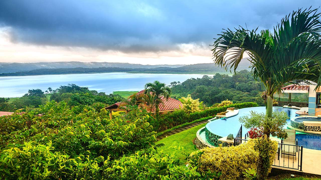 A pool with trees and a house on a hill facing a body of water under cloudy skies