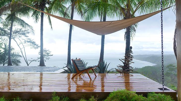 A chair below a hammock on a wet wooden platform overlooking palm trees and the sea