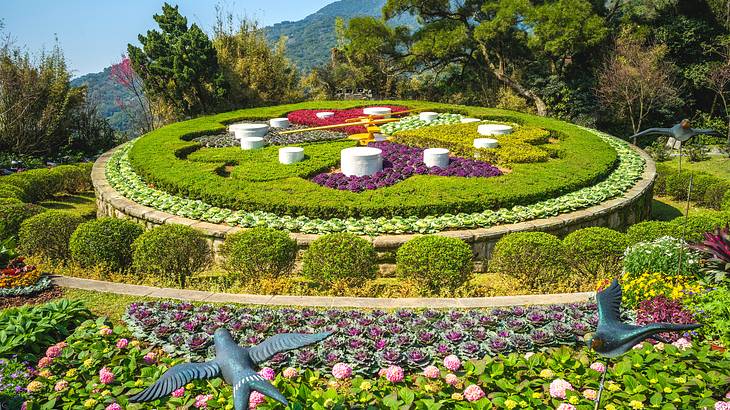 A round flower clock surrounded by bushes, flowers, and trees, with mountains behind