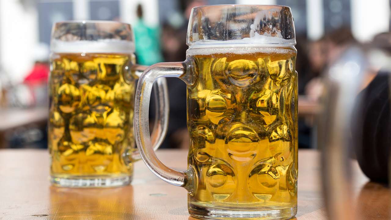 Two stein glasses full of beer on a wooden surface against an out-of-focus background