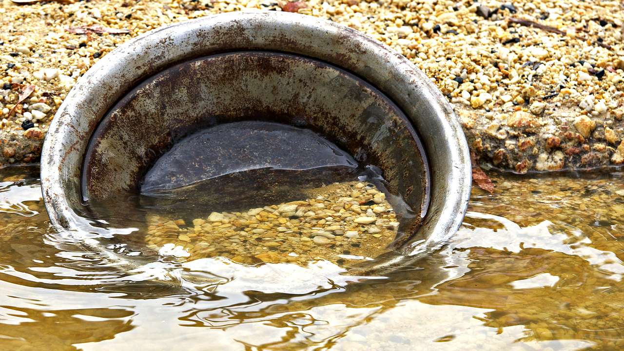 A metal pan with sand and water inside it on the gravelly banks of a river