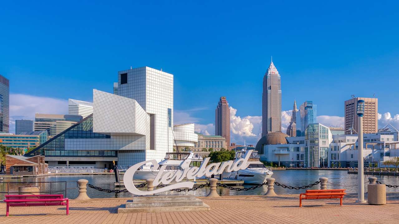 A path with benches and a "Cleveland" sign next to a waterway and city skyline