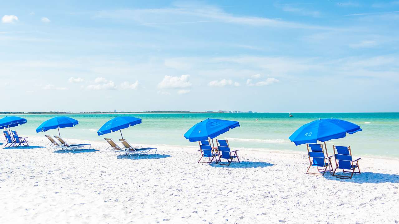 A white, sandy beach with blue umbrellas lining the green ocean in the distance
