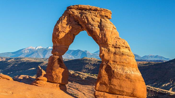 A red sandstone arch in a park with mountains under a blue sky