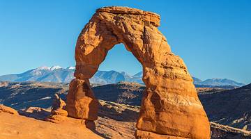 A red sandstone arch in a park with mountains under a blue sky