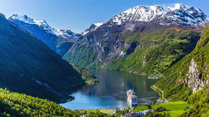 A fjord with a cruise ship on it surrounded by tall mountains with snow and greenery