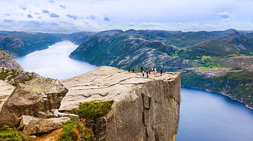 A group of people on a clifftop next to a body of water and green hills