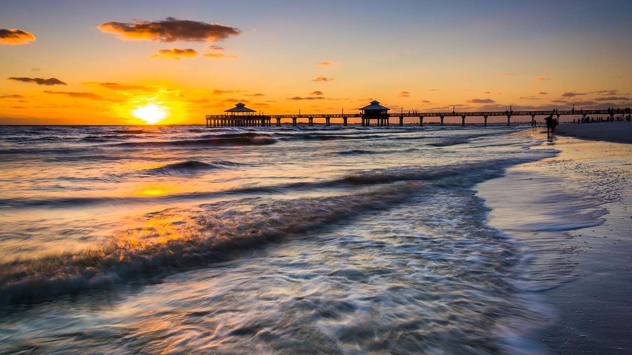 Water flowing towards a beach in front of a pier boardwalk against a sunset sky