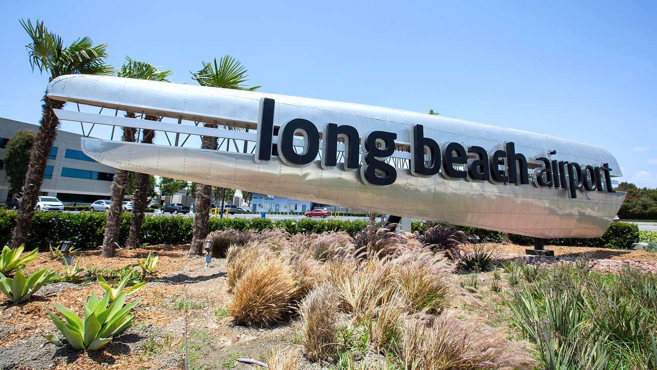 A silver metal sign that says "long beach airport" next to shrubbery and palm trees