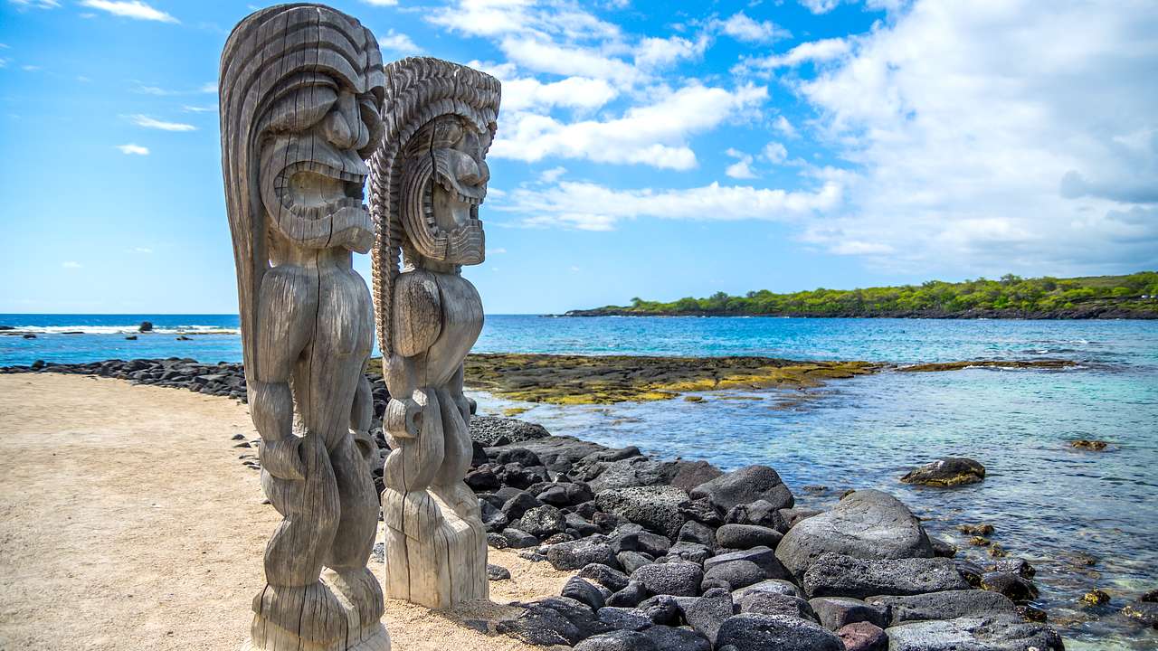 Tall wooden carvings on a rocky beach with a partly cloudy blue sky in the background
