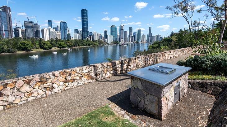 A pathway in a park with a barbecue area overlooking water and buildings at the back