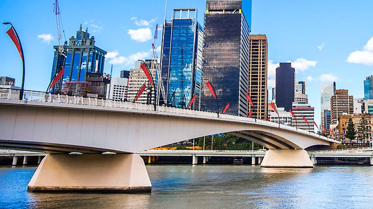 A bridge with red flags on the left with tall city buildings at the back by a river A bridge with red flags on the left with tall city buildings at the back by a river