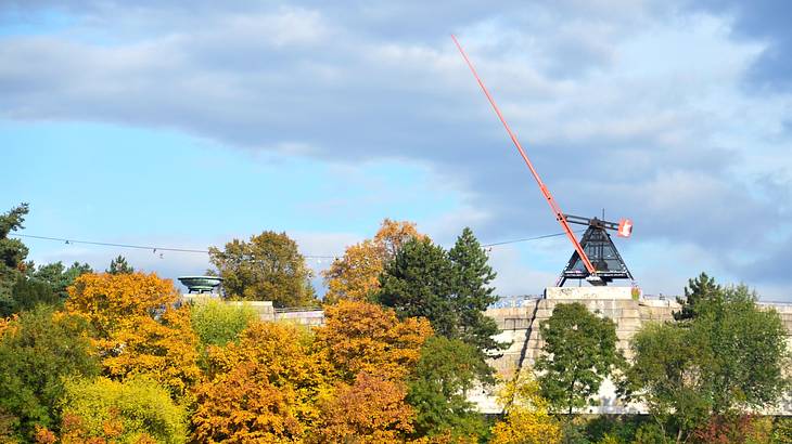 A giant metronome on a brick platform behind a row of trees in autumn colors