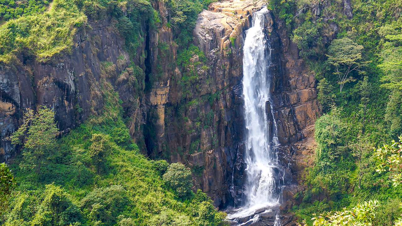 A tall thin waterfall falling down a cliff surrounded by greenery from far away