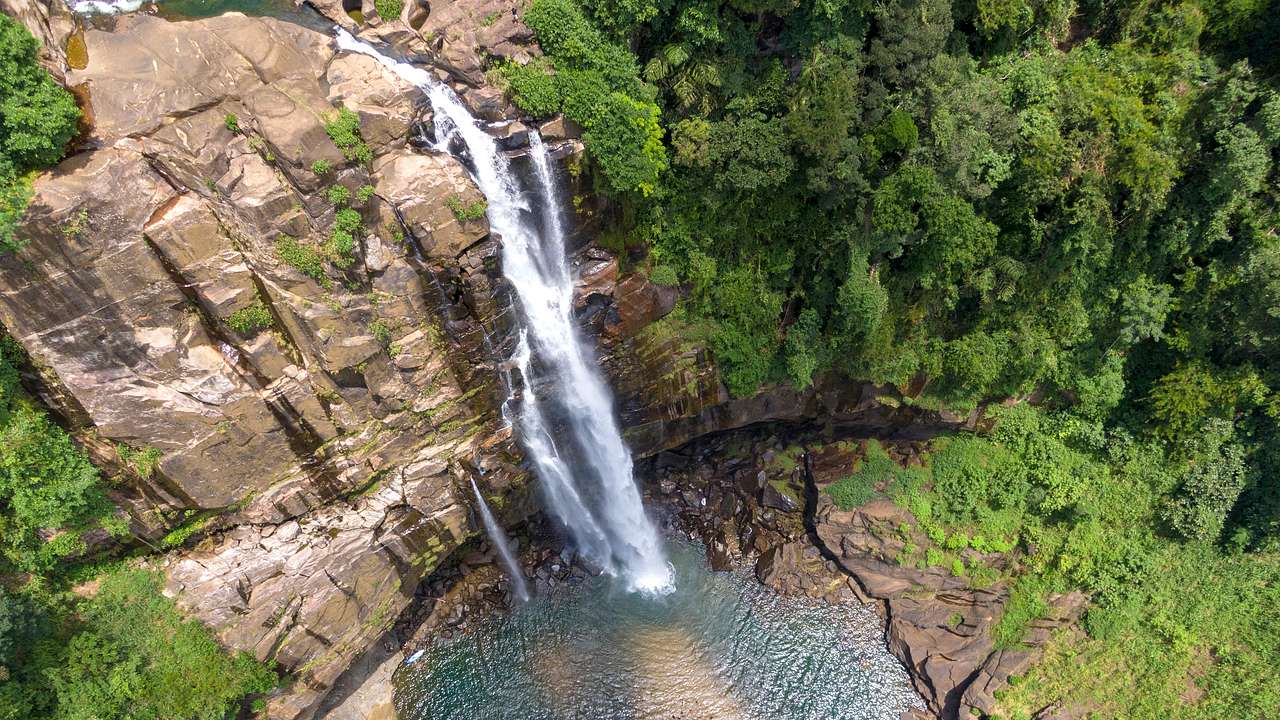 A tall and thin waterfall falling off rock into water below with jungle around
