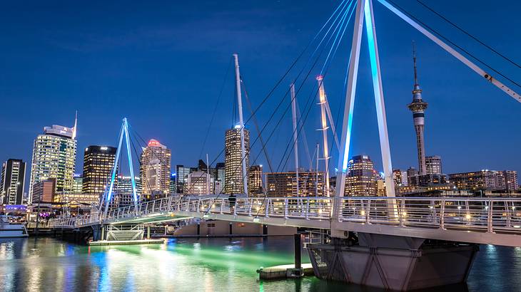 Lit-up buildings in a city facing a body of water, boats, and a bridge at night