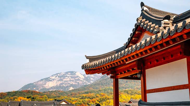 The top red roof of a temple with trees, mountains and blue sky at the back