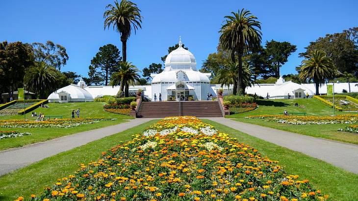 A white conservatory surrounded by grass, flowers, and palm trees on a clear day