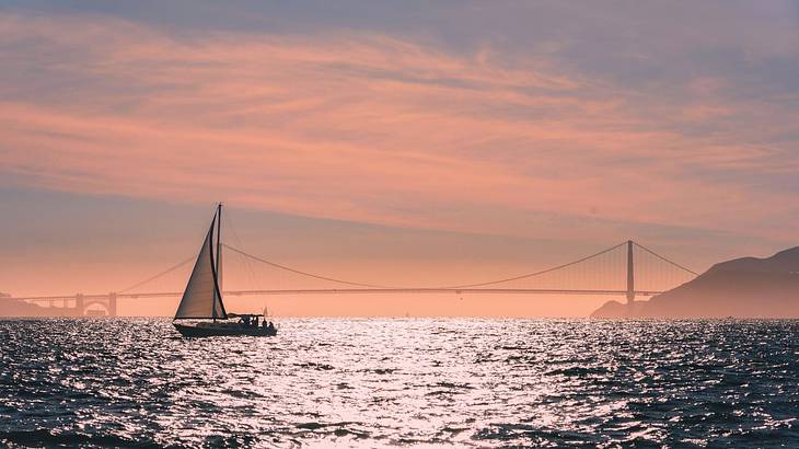 A sailboat on the water with a suspension bridge in the distance at sunset A sailboat on the water with a suspension bridge in the distance at sunset