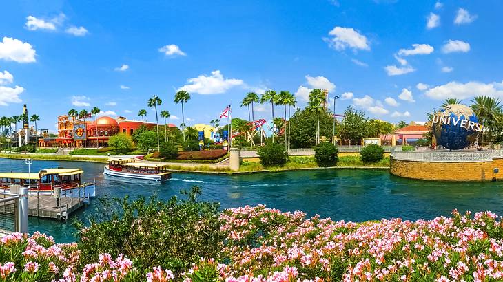 A body of water with flowery bushes in front and colorful structures and trees behind