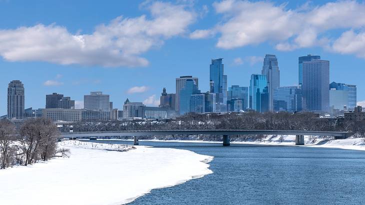 A river next to an area of snow, bare winter trees, and a city skyline