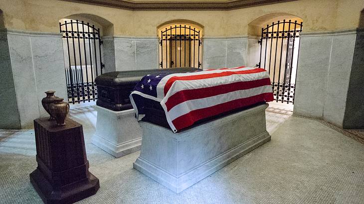 Two caskets, one covered in a US flag, in a stone room with iron gates