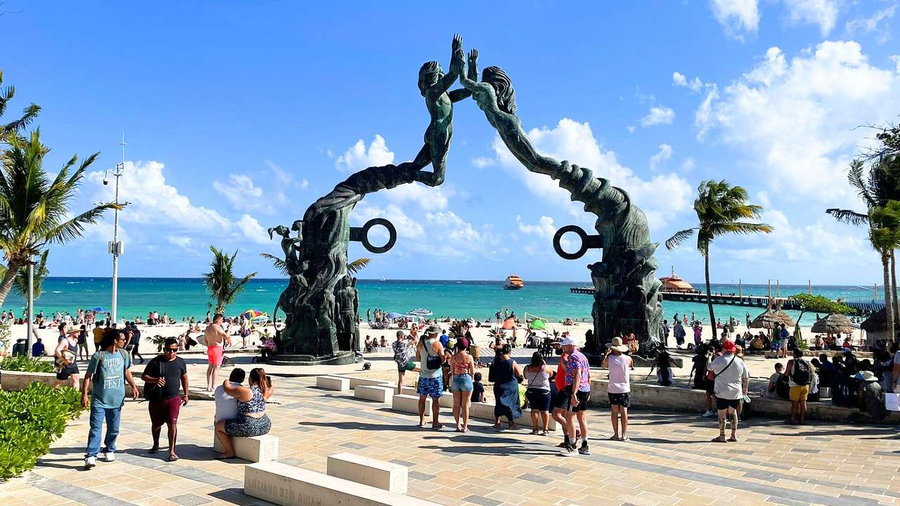 Two intertwined statues in front of the ocean and a beach with people walking around