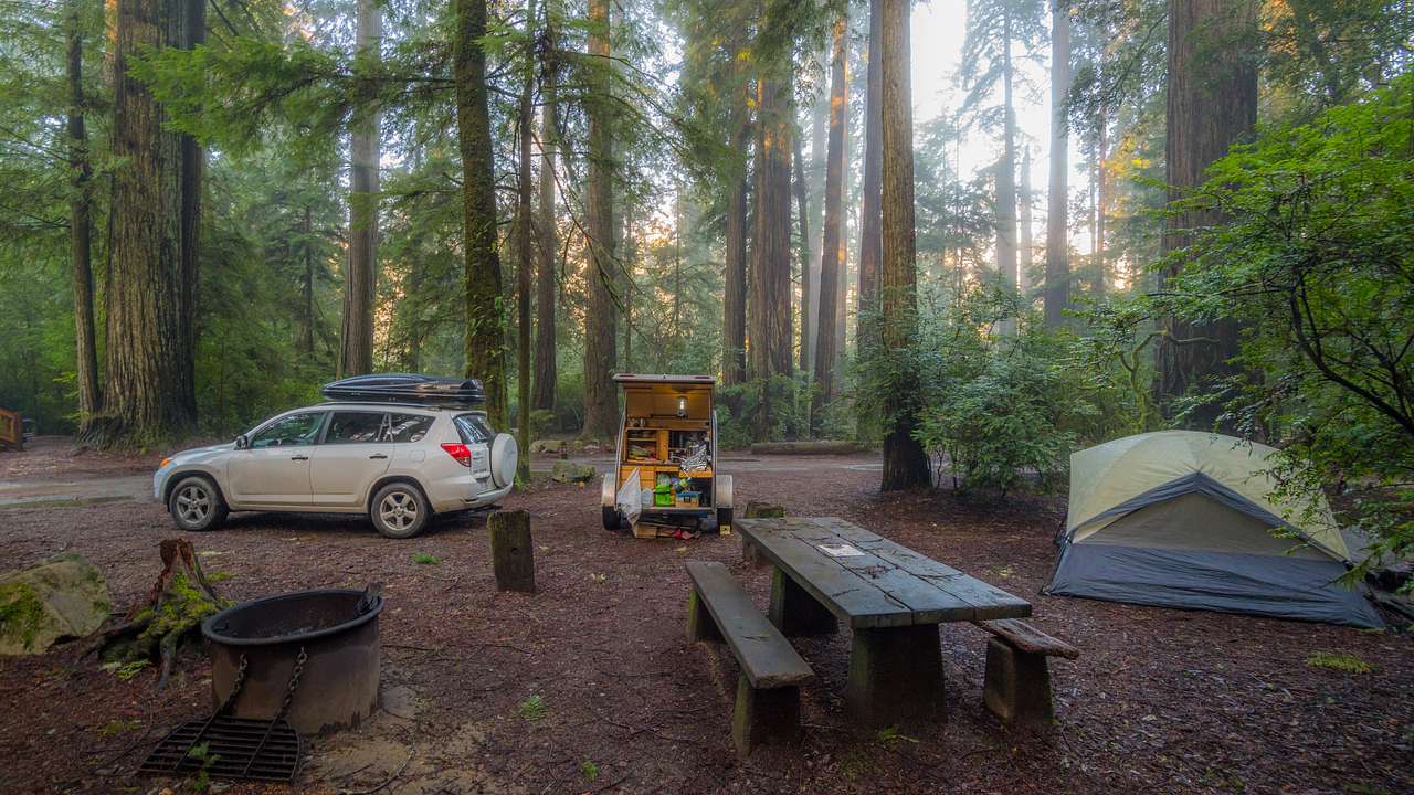 A camping site with a tent, a bench, and a van in the middle of a forest