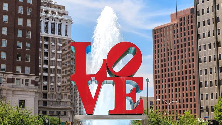 A red "LOVE" sculpture next to a fountain, trees, and city buildings