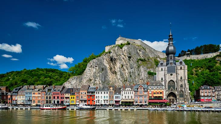 A hill with greenery next to small colorful buildings by a river on a sunny day