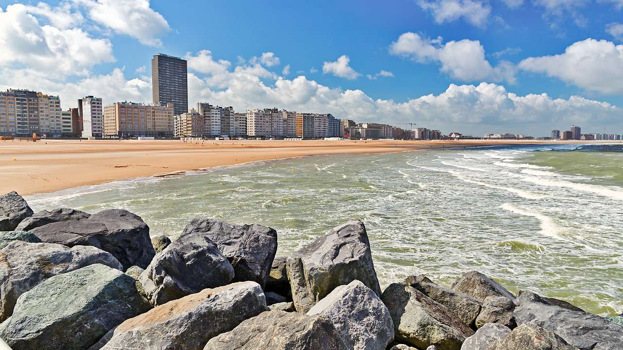 A sandy beach and the ocean next to buildings with rocks in the foreground