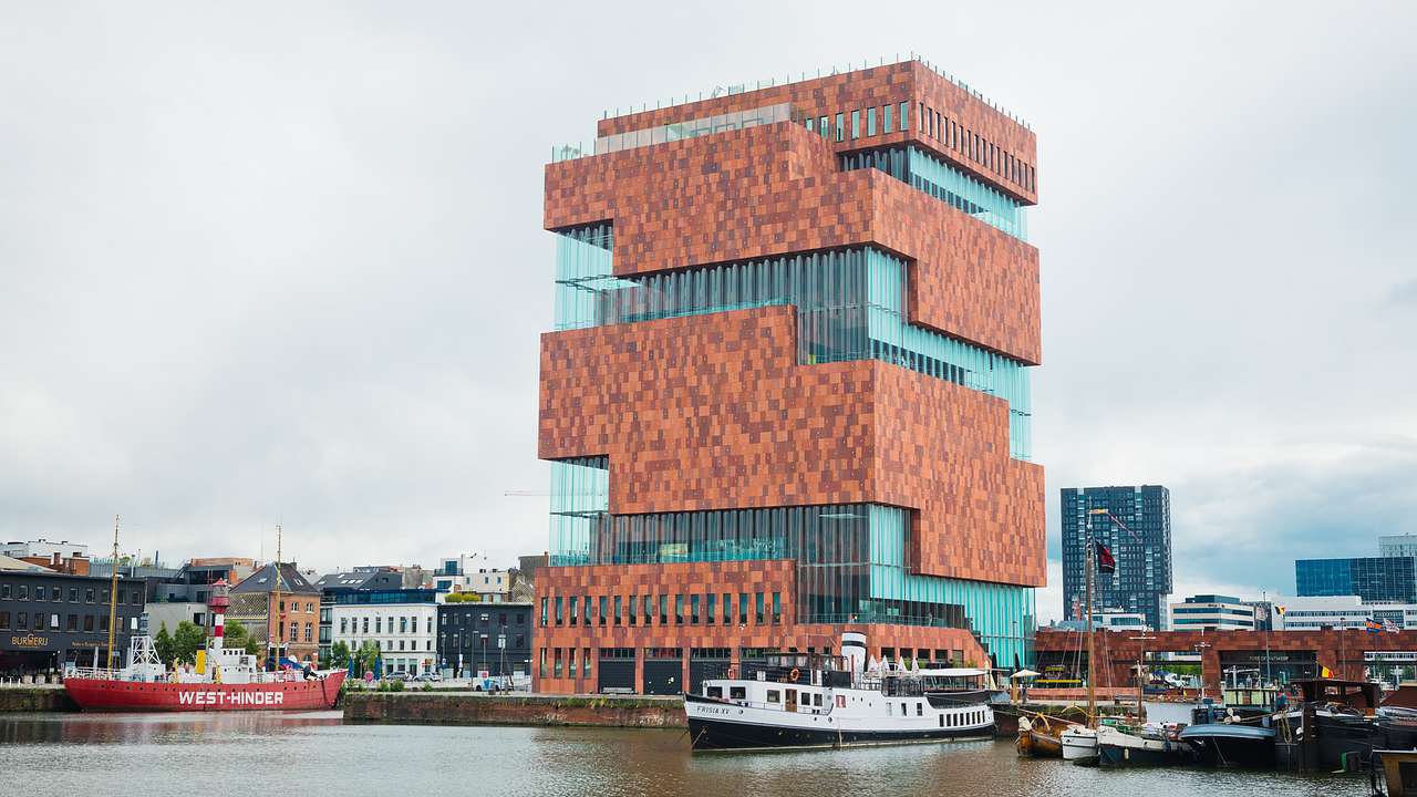 A red brick and glass structure along the river with a yacht in front on a cloudy day