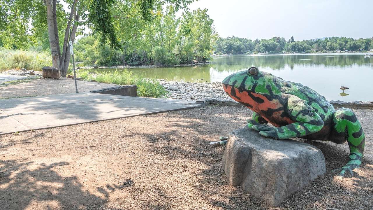 A green and red frog sculpture next to trees and a lake on a bright day