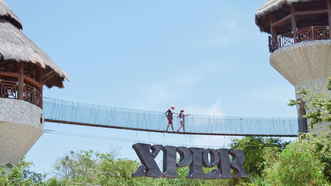 Two people walking over a suspension bridge with a sign that says "Xplor" below it