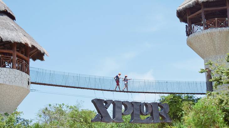 Two people walking over a suspension bridge with a sign that says "Xplor" below it